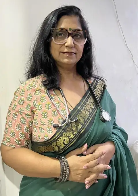 A woman in a dark green saree with a gold-bordered pallu over a peach block-printed blouse, wearing tortoiseshell glasses and a stethoscope round her neck, standing with her hands clasped against a plain pale wall.