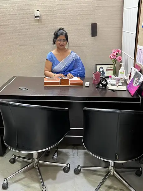A woman in a blue saree seated behind a large dark wooden desk in the consultation room, two black swivel chairs facing her across it, and a framed name card, a vase of pink flowers and a bottle of hand sanitiser at one end of the desk.